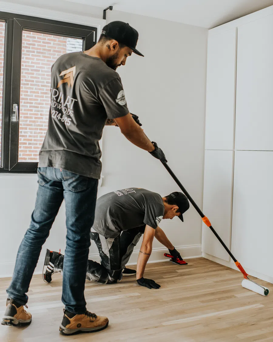 Two workers cleaning and polishing light wooden floors in a bright room with white walls and cabinets.