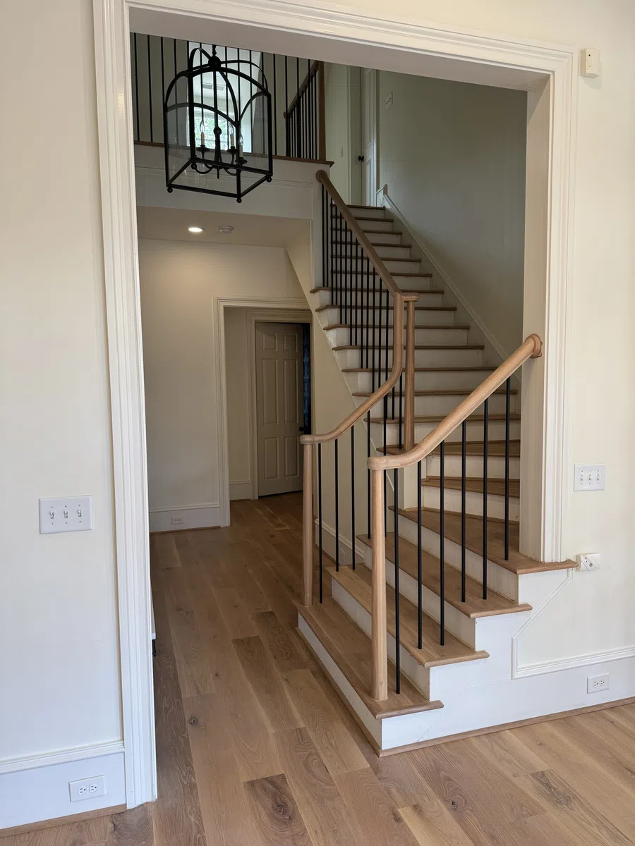 Modern wooden staircase with black metal balusters and light hardwood flooring in a neutral colored hallway.