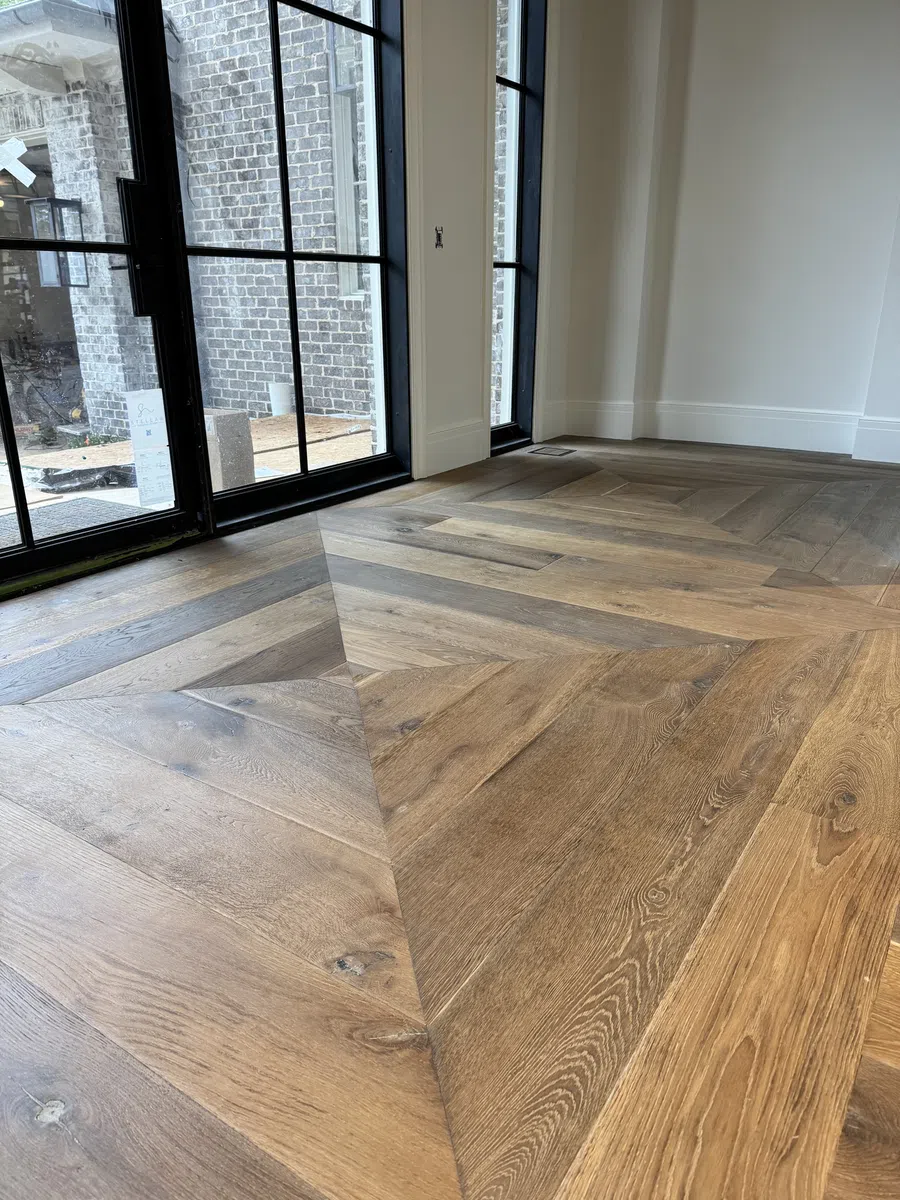 Chevron-patterned wooden flooring near large black-framed windows in a modern bright room with white walls.