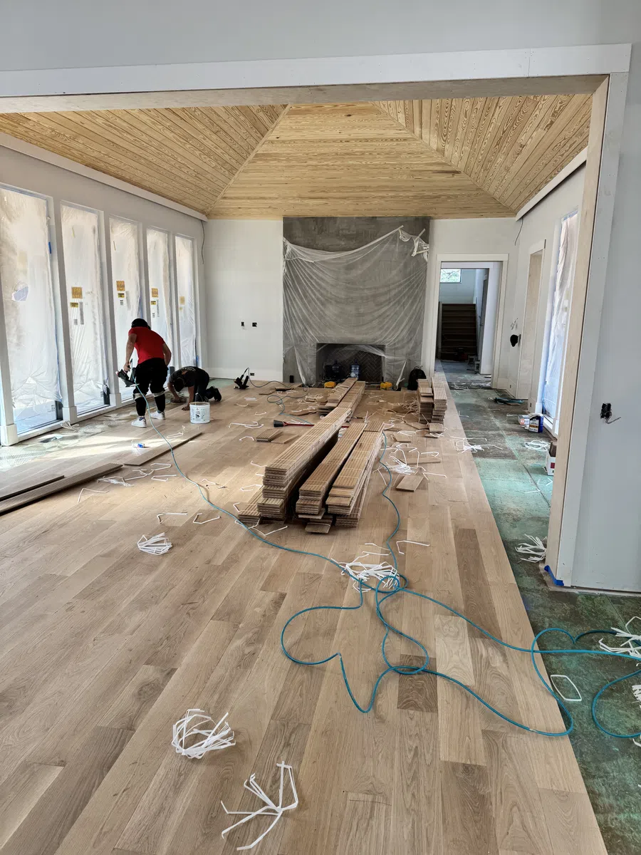 Workers installing hardwood flooring in a room with high wooden ceilings and large windows covered for renovation.