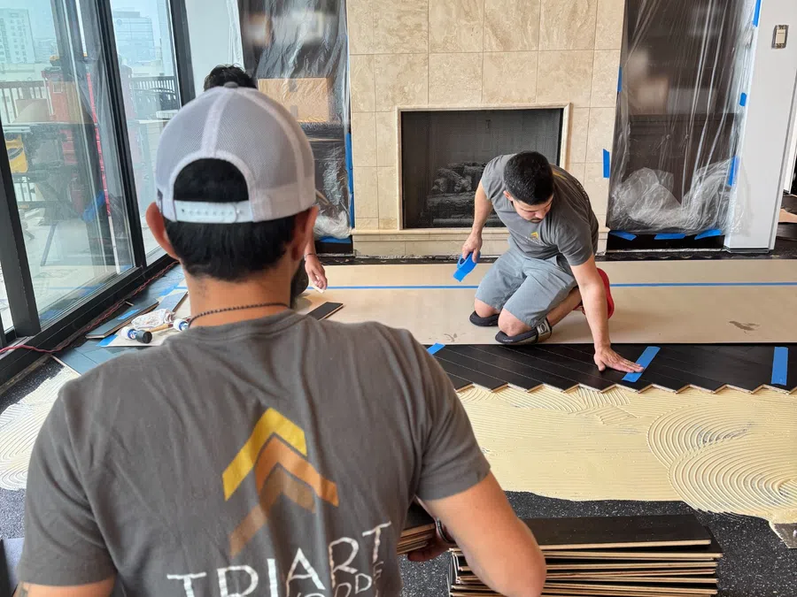 Two workers installing dark hardwood flooring in a modern living room with a fireplace and large windows.