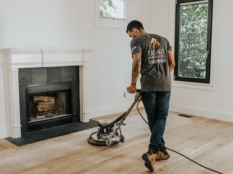 Man sanding hardwood floor near fireplace in bright room with large window and white walls
