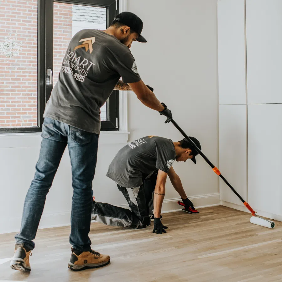 Two workers installing or finishing a hardwood floor in a bright, modern room with large window and white walls.