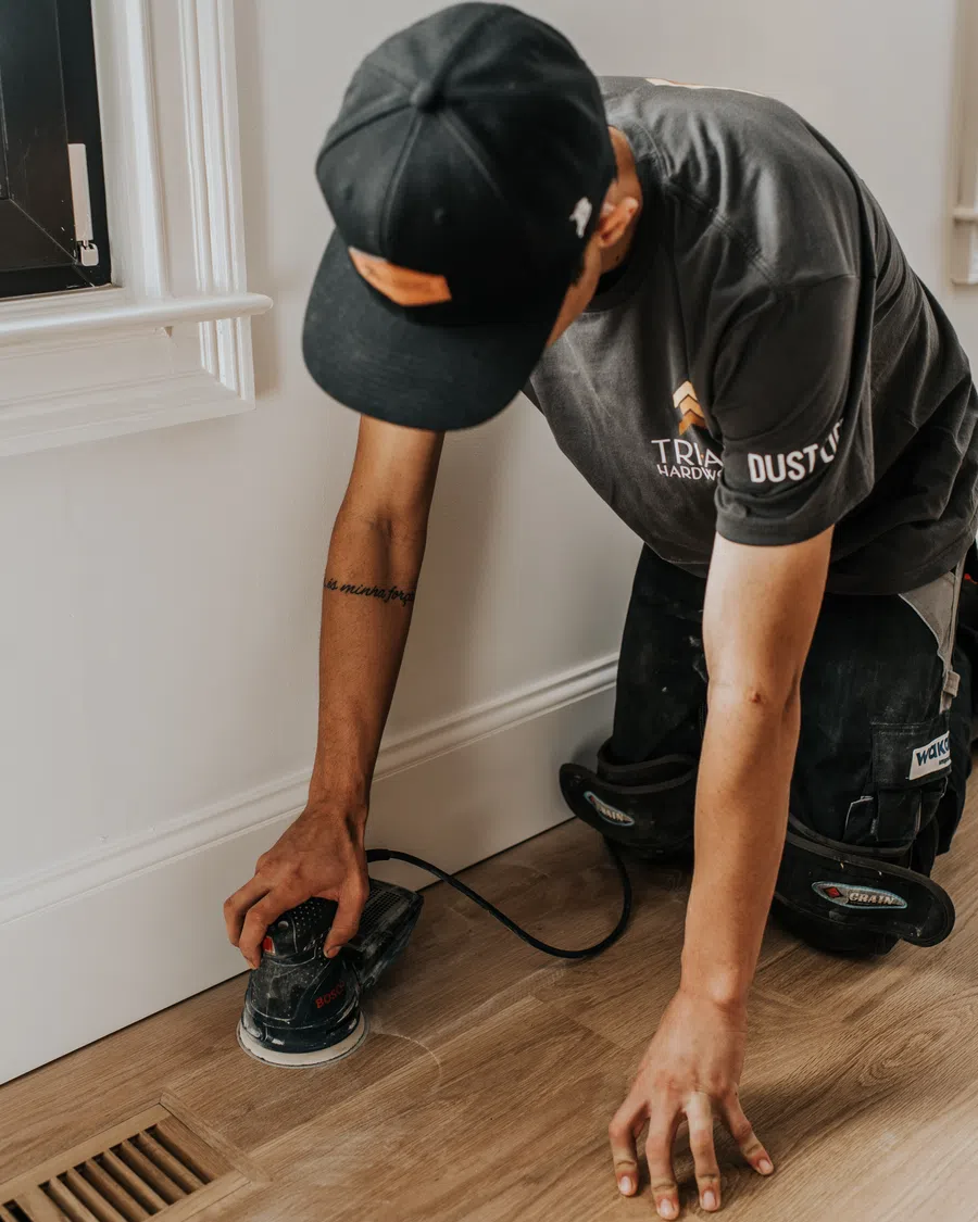 Worker wearing black cap and shirt sanding wooden floor near white wall with power sander