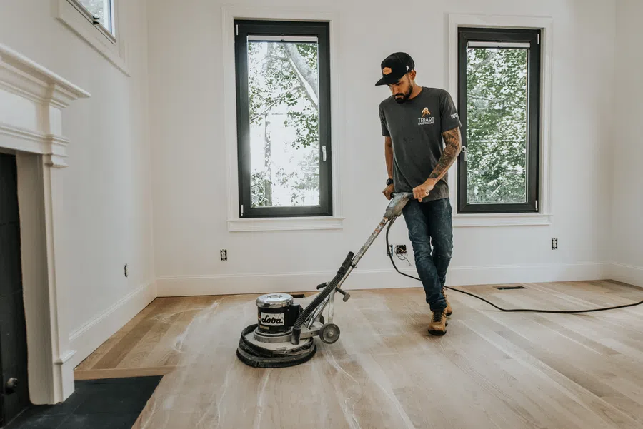 Man refinishing hardwood floor with floor buffer machine in bright room with two windows.