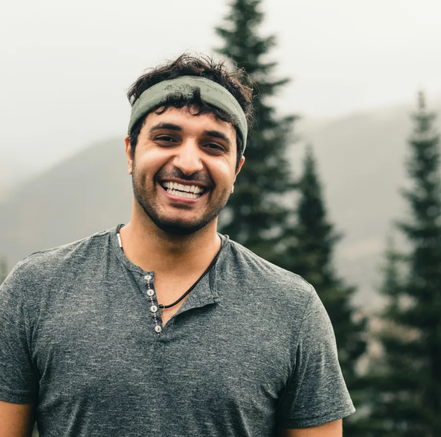 Smiling man wearing a headband and grey shirt stands outdoors with evergreen trees and mountains in the background