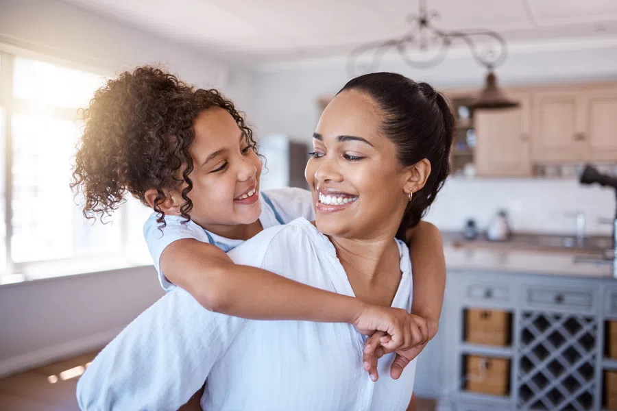 Smiling mother and daughter embracing joyfully in a bright, modern kitchen with natural sunlight.