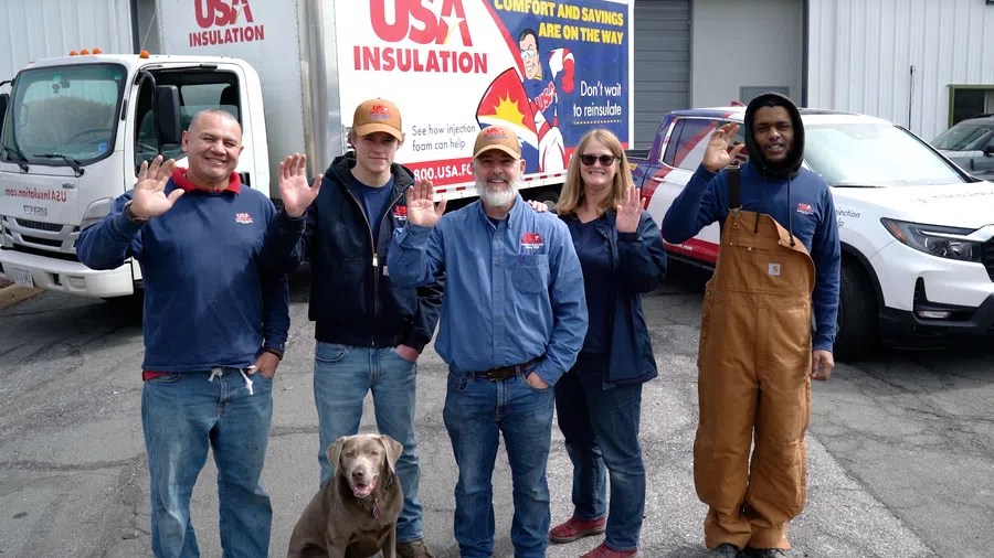 Five people and a dog stand smiling and waving in front of USA Insulation trucks outside a building.
