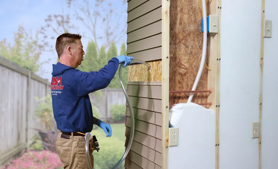 Technician in blue jacket installing insulation inside home wall with siding outdoors on a sunny day