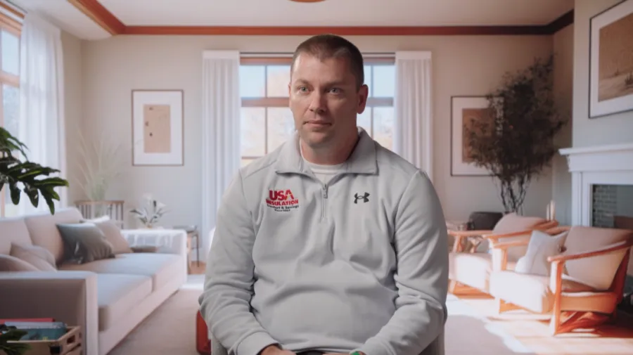 Man in white USA Insulation jacket sitting in a bright, modern living room with beige and white decor.