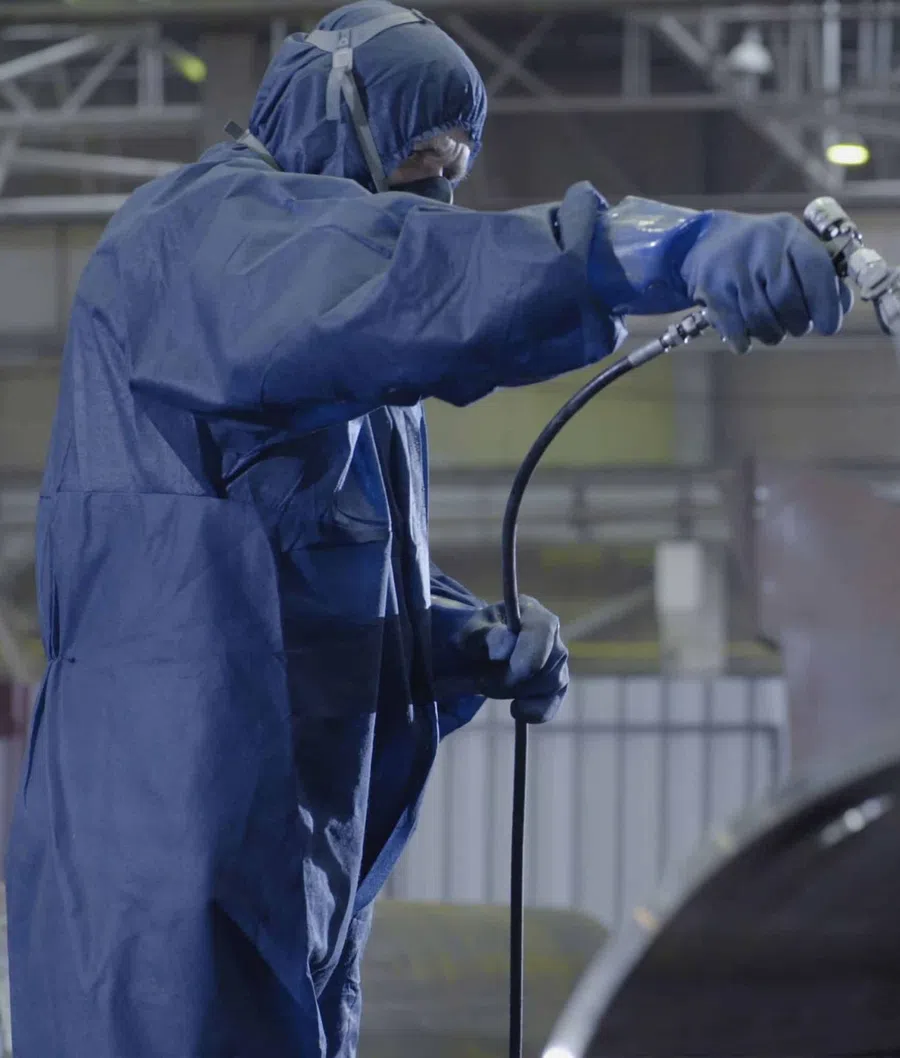 Worker in protective suit spray painting industrial equipment inside factory environment.
