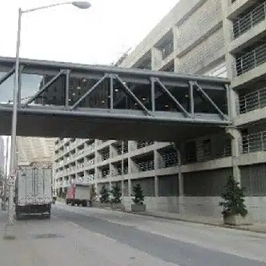 Urban street scene with a pedestrian bridge connecting two buildings over a road with trucks and trees.
