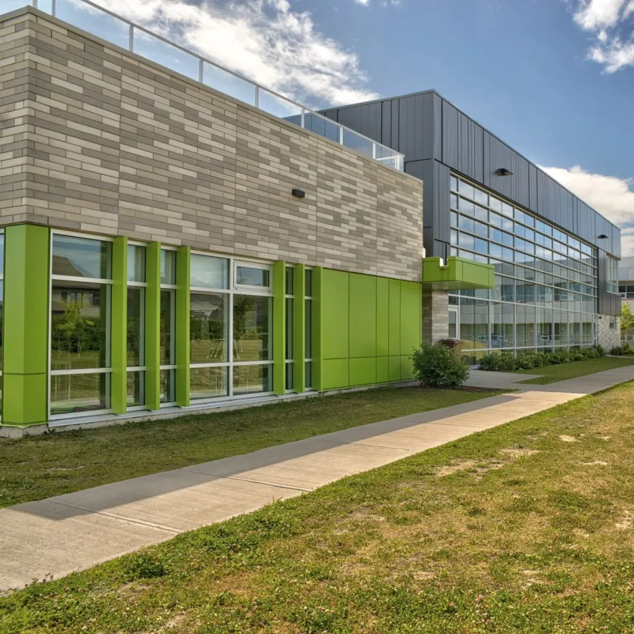 Modern school building with green accents, large windows, grassy lawn, and clear blue sky with clouds.