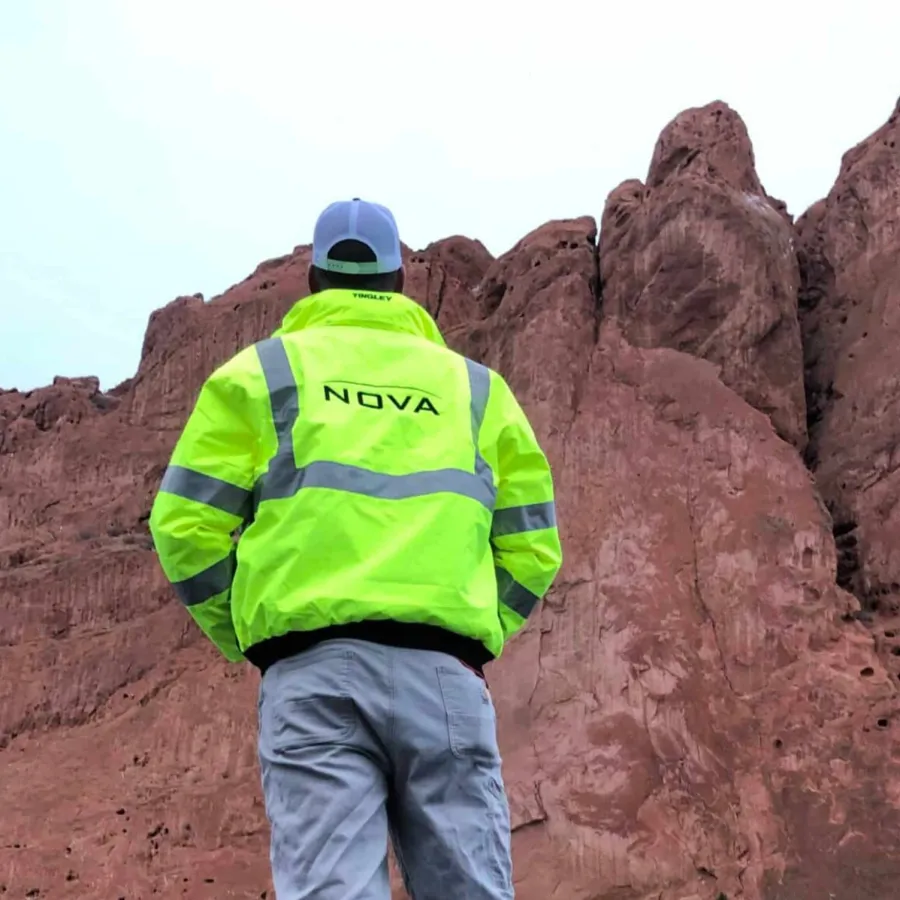 Person wearing a bright yellow reflective jacket with NOVA logo standing in front of tall red rock formations.