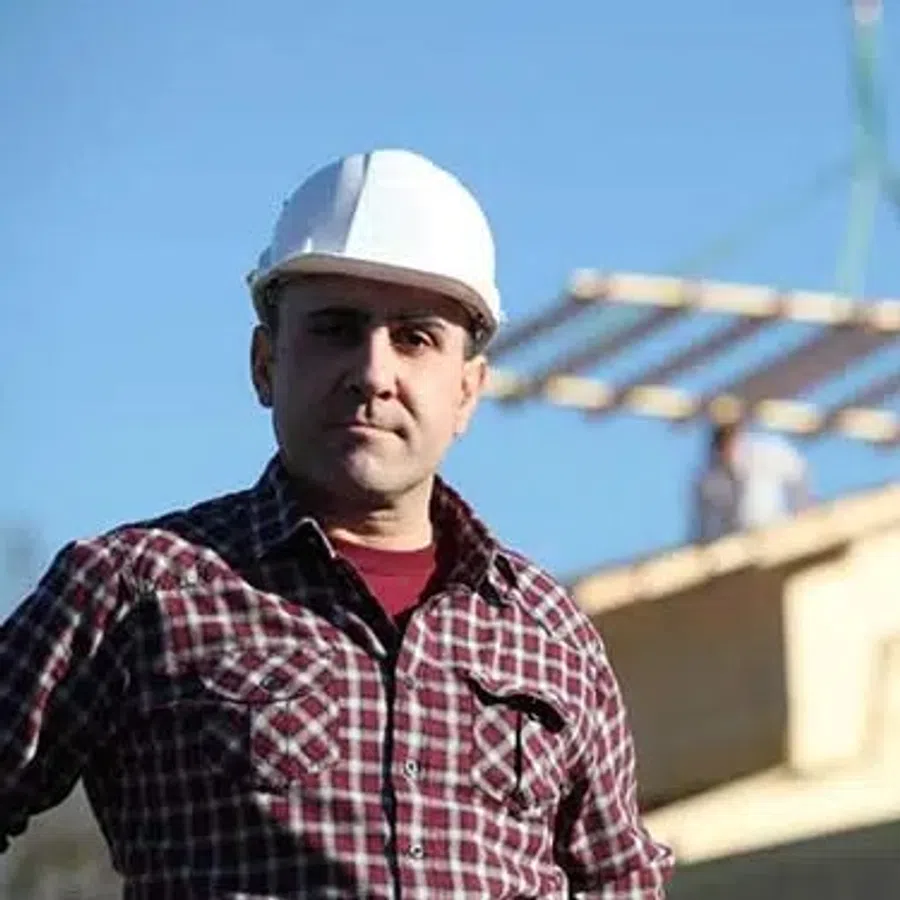 Construction worker wearing a white helmet and plaid shirt with building materials lifted by crane in background