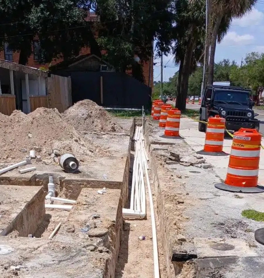 Construction site with trench, pipes, orange barricades, dirt piles, and nearby parked vehicles on a sunny day.