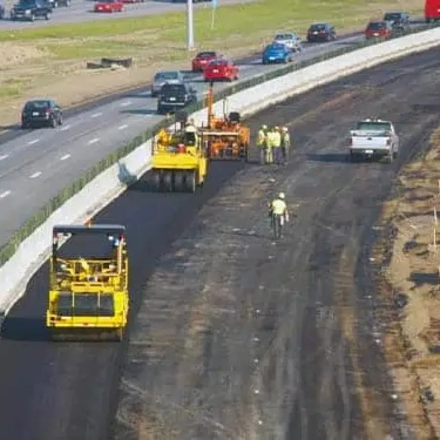 Construction workers and machinery paving a new highway lane next to an active multi-lane road.