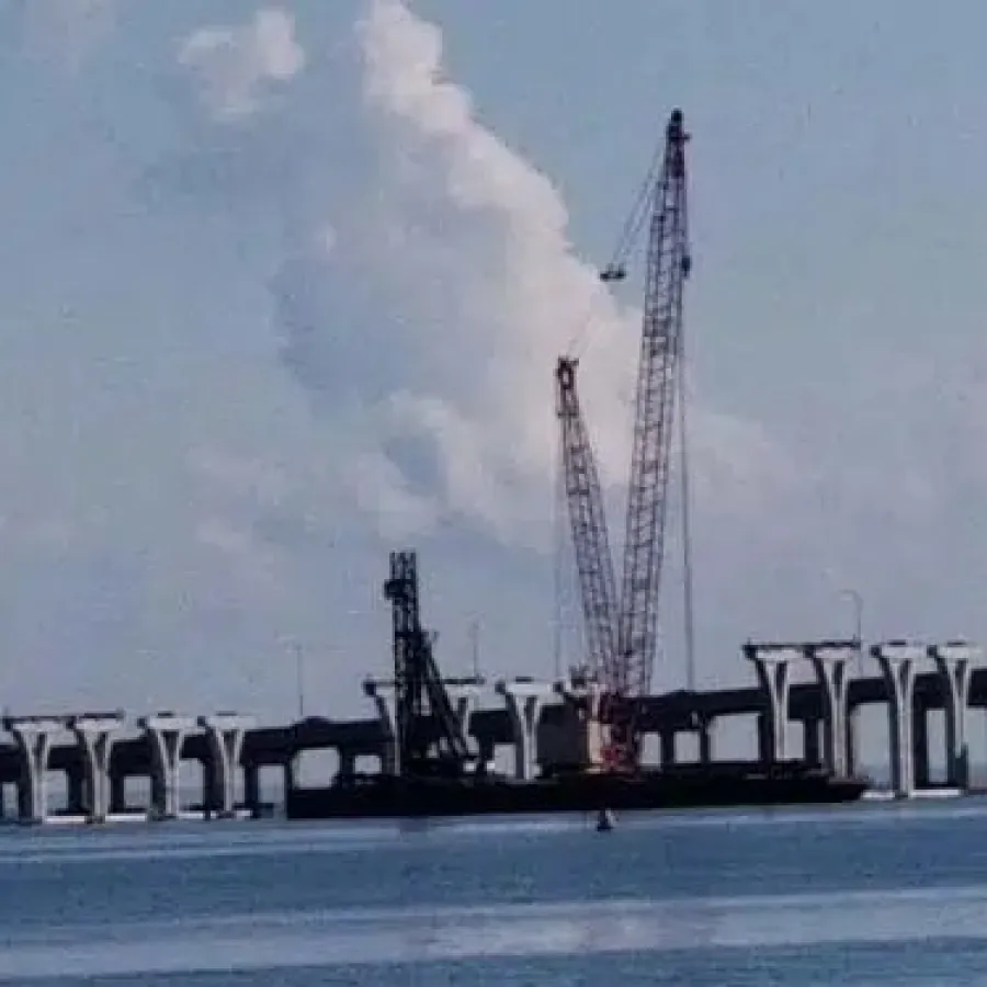 Construction cranes on a barge working on a large bridge over calm water under a partly cloudy sky.