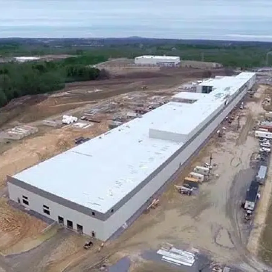 Aerial view of a large industrial warehouse under construction surrounded by dirt roads and forested area.