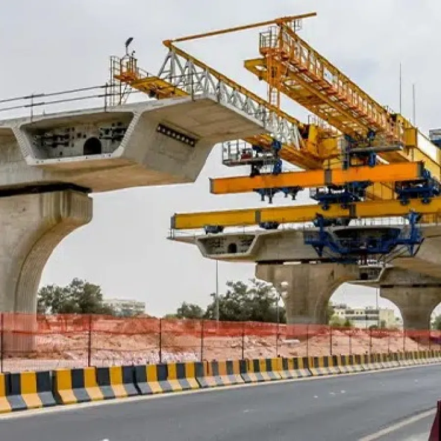 Concrete bridge segments being assembled using heavy yellow construction cranes over a highway with safety barriers.
