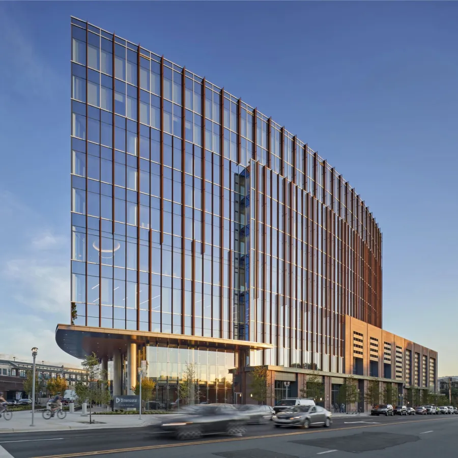 Modern glass office building with curved facade reflecting sunlight and vehicles on busy street in front.
