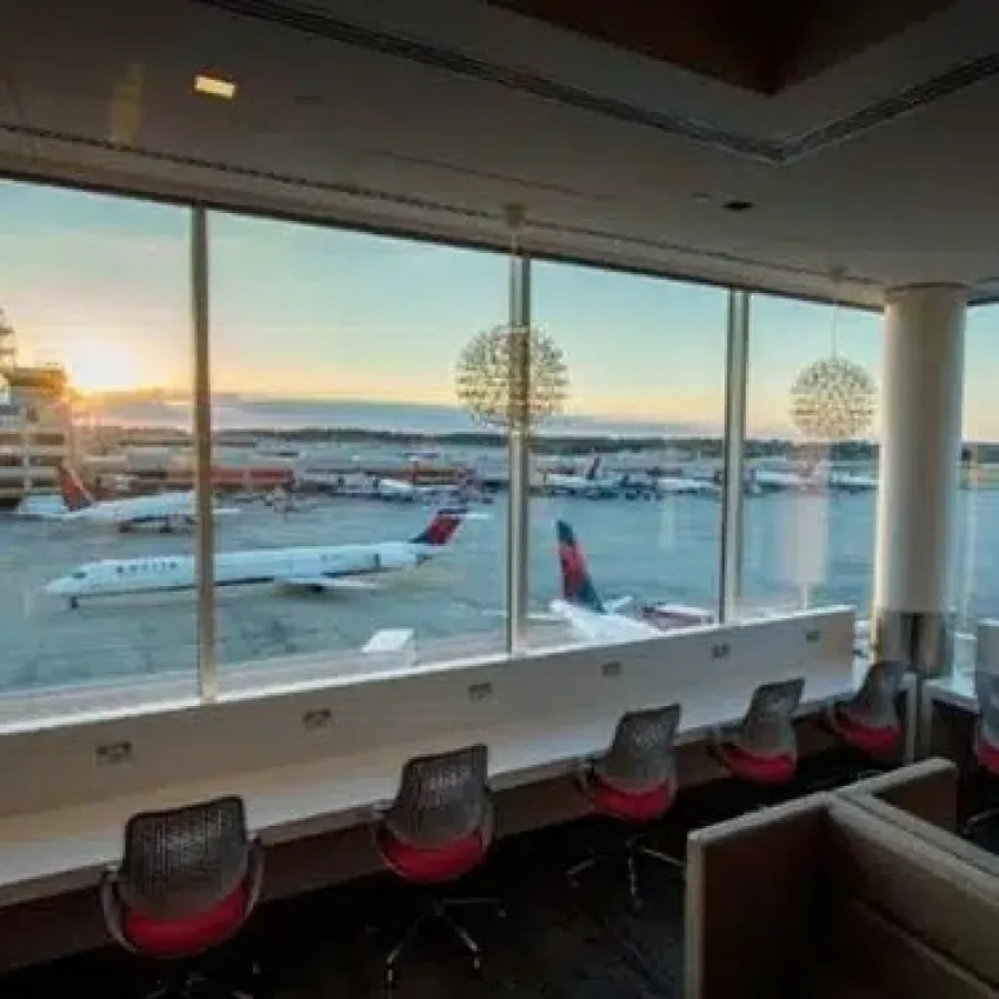 Airport lounge workspace with desks, chairs, large windows overlooking airplanes and runway at sunset.