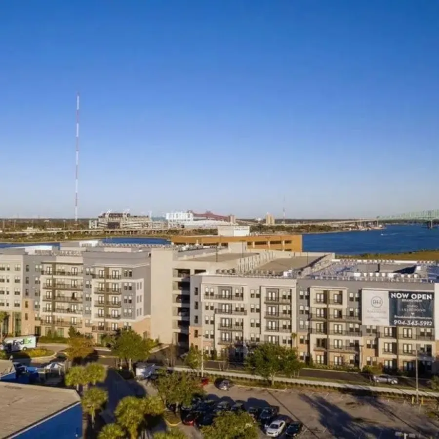 Modern apartment complex by the river with clear blue sky and a green bridge in the background.