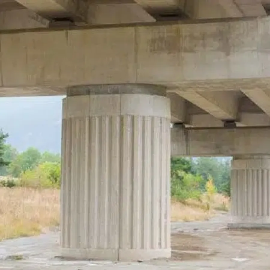 View of large concrete bridge pillars supporting an elevated roadway over a natural landscape with trees and grass.