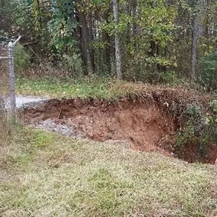 Large erosion hole exposing red soil near fence and forest with trees and green vegetation.