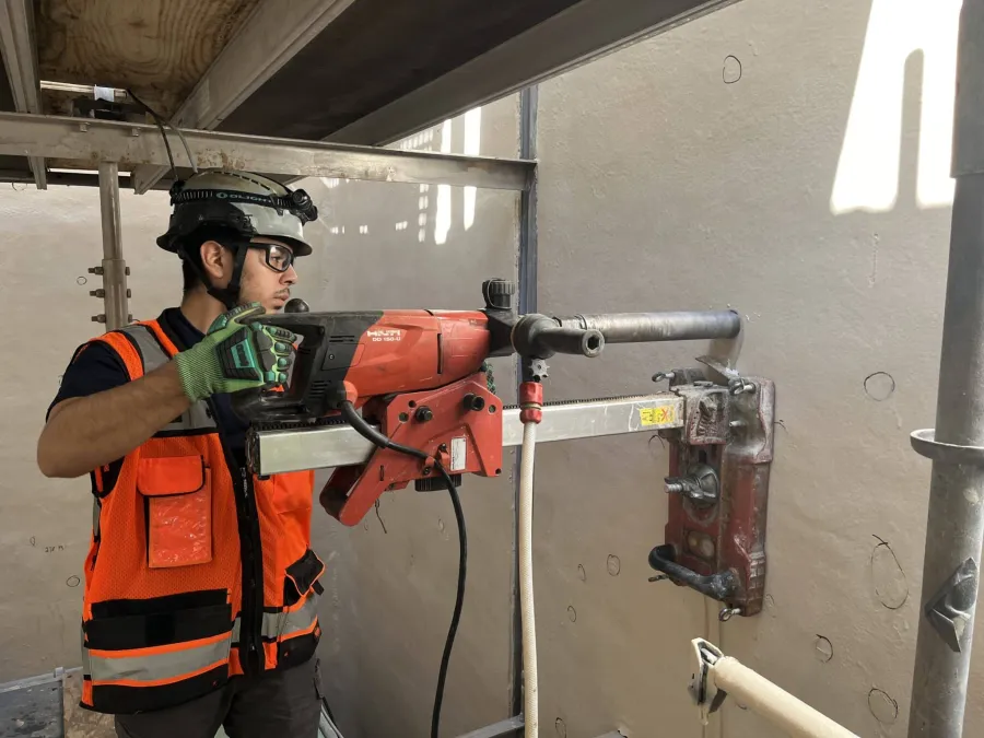 Construction worker in safety gear operating a large drilling machine mounted on a wall indoors.