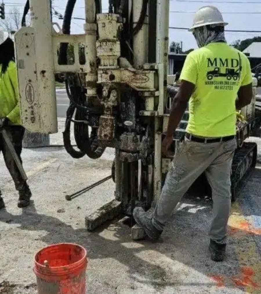 Construction workers operating drilling machinery on a street wearing neon yellow safety shirts and helmets.