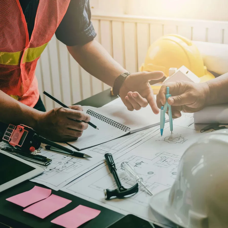 Two construction engineers discussing blueprints with drawing tools and safety helmets on table.