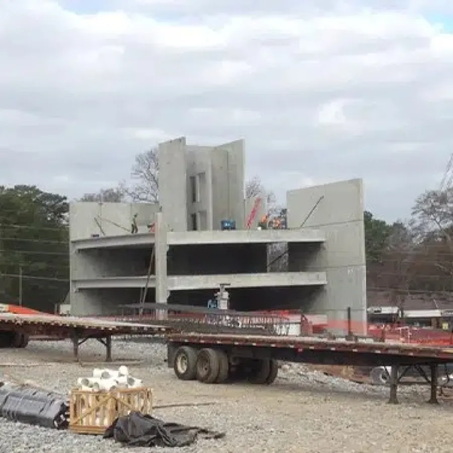 Concrete building under construction with cranes, flatbed trailer, and construction materials at site.