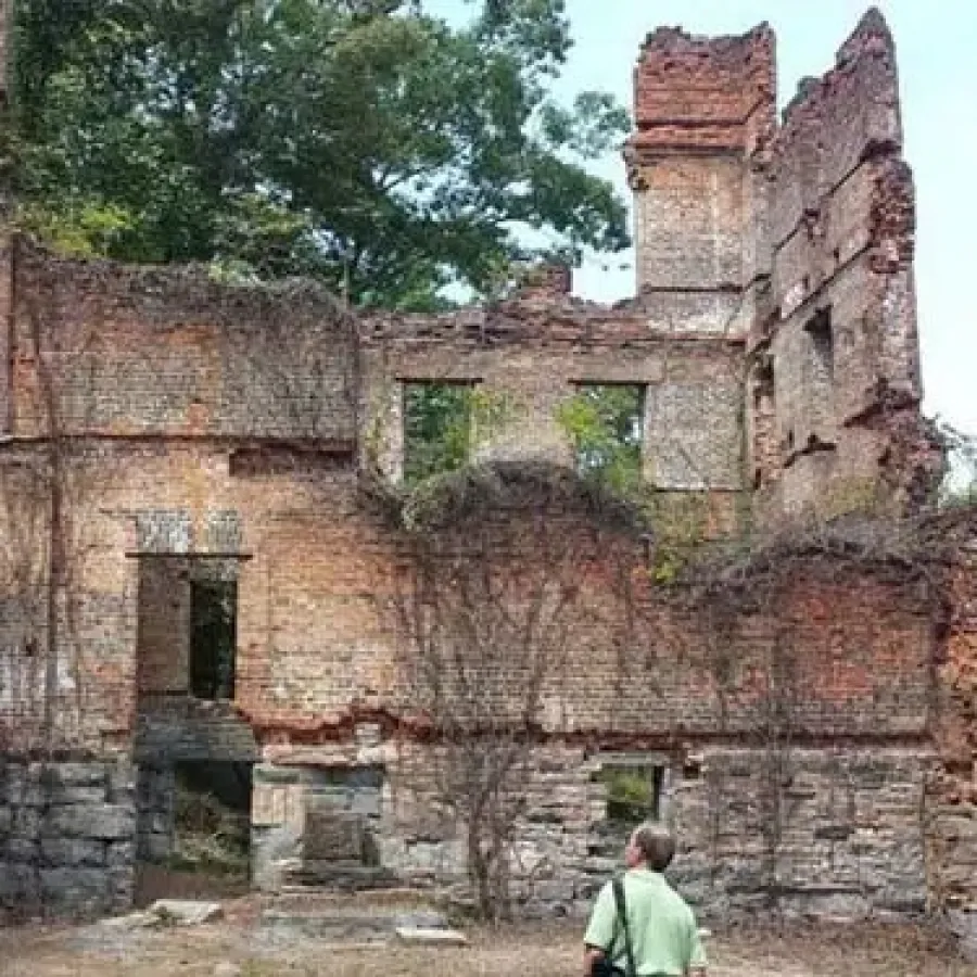 Person standing inside a large, abandoned brick building ruin with overgrown plants and empty window frames.