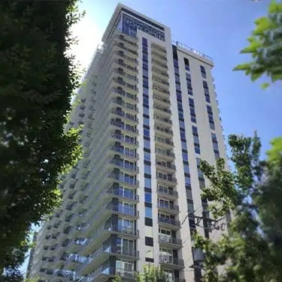 Modern high-rise apartment building with balconies surrounded by trees under a clear blue sky.