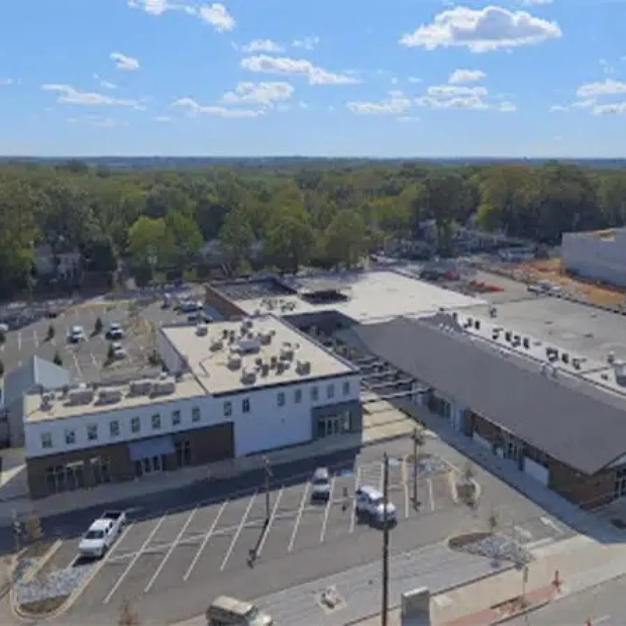 Aerial view of a commercial complex with parking lots, trees, and a construction site under a blue sky.