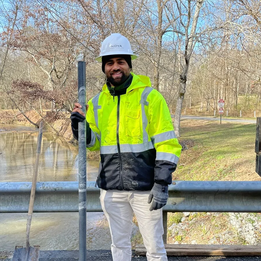Smiling construction worker in neon yellow jacket and hard hat holding a metal rod near a river and guardrail.