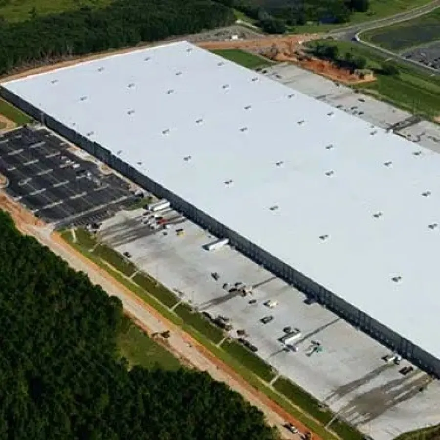 Aerial view of a large industrial warehouse surrounded by parking lots and greenery under clear skies.