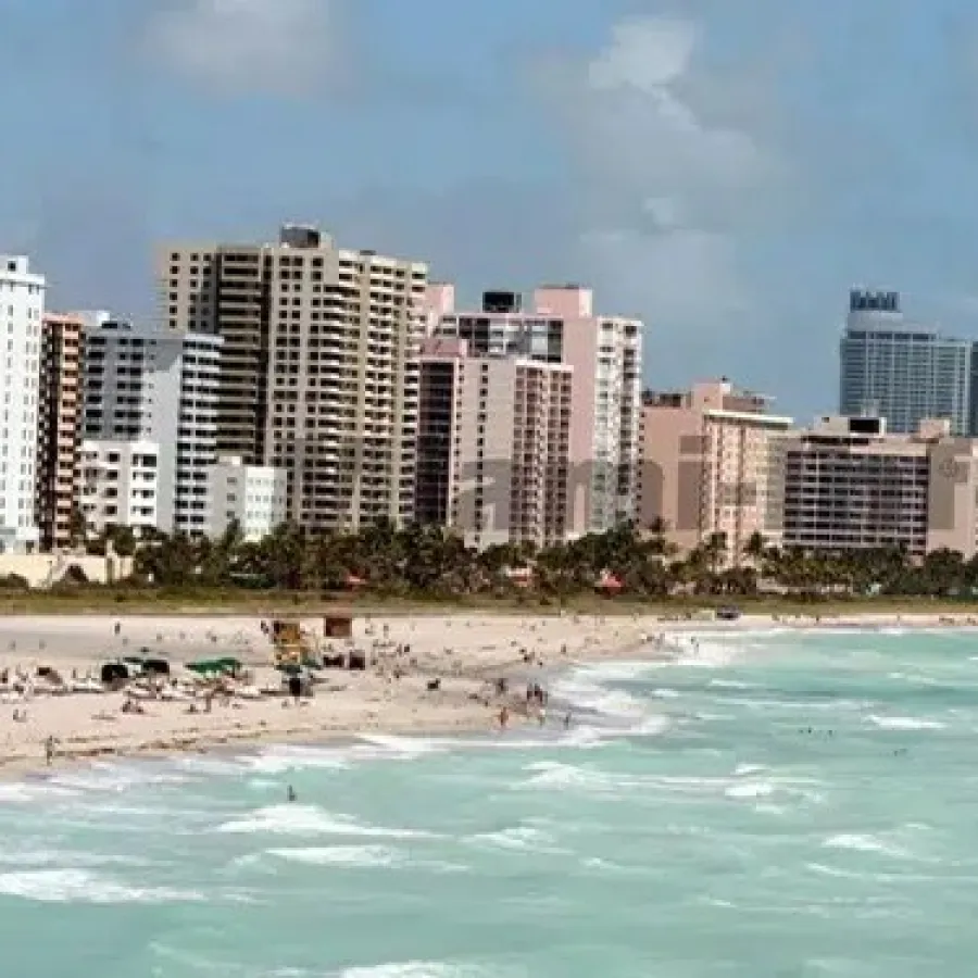 Sunny beach with turquoise water and waves, lined with high-rise hotels and palm trees under a partly cloudy sky.