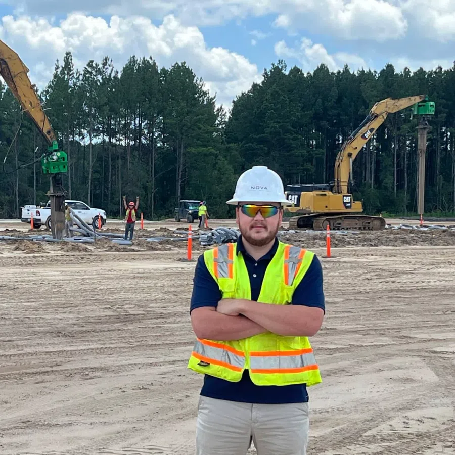 Construction worker in safety vest and helmet stands on site with excavators and forest background under blue sky.
