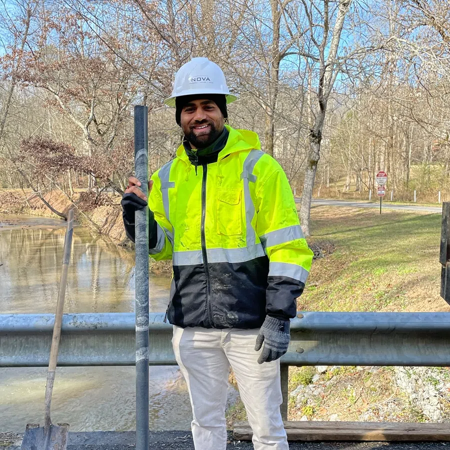 Smiling construction worker in neon yellow jacket and hard hat holding a metal rod near a river and guardrail.