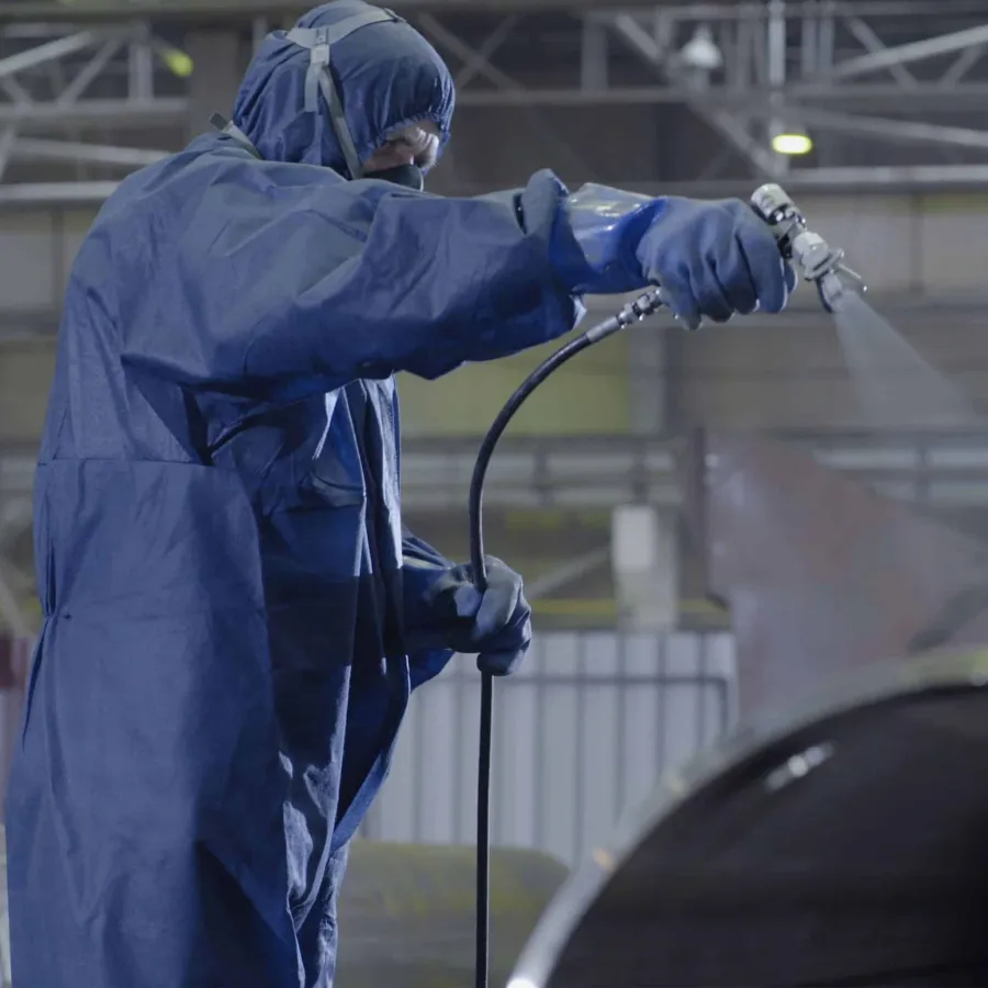 Worker in protective suit spray painting industrial equipment inside factory environment.