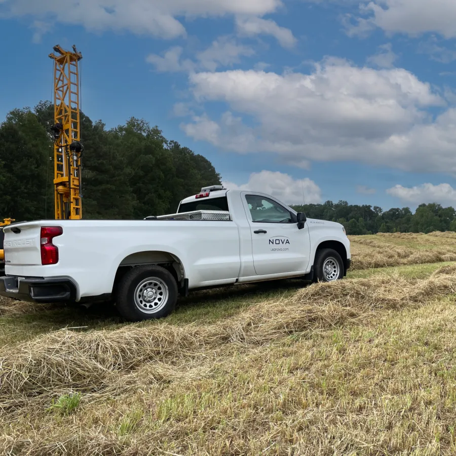 White NOVA pickup truck parked on a hay field with a drilling rig and trees under a partly cloudy sky.