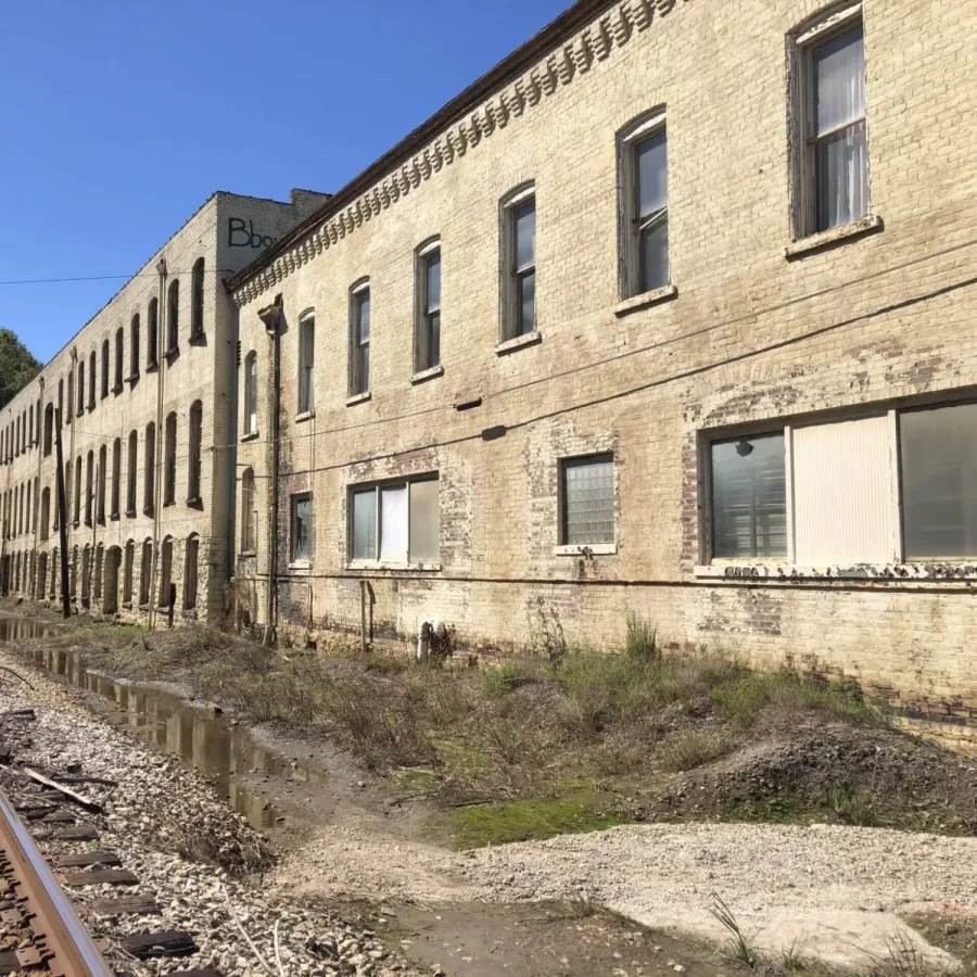 Old industrial building next to railroad tracks with trees and clear blue sky in background.