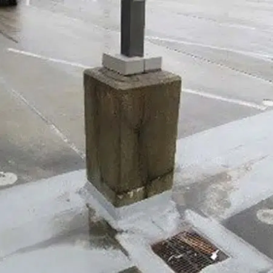 Concrete pillar base in a parking lot showing wet patches and a nearby storm drain on asphalt surface.