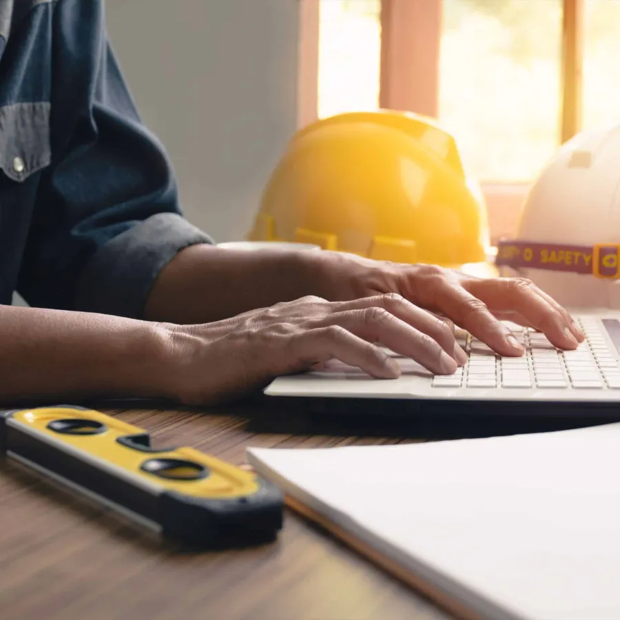 Hands typing on laptop with construction helmets and level tool on desk by window light