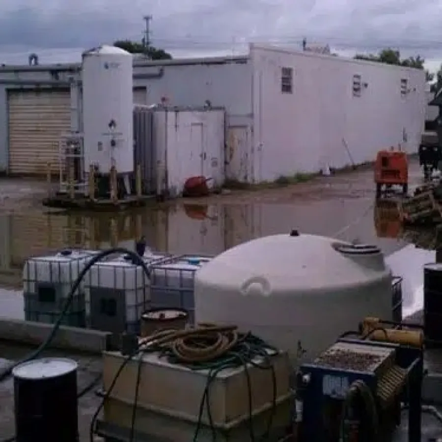 Industrial storage tanks and barrels near a flooded yard with white buildings under cloudy skies
