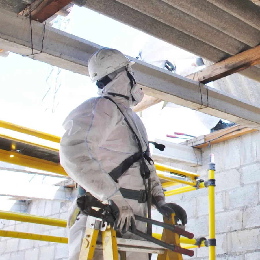 Two workers in protective suits and helmets carry corrugated metal sheets at a demolition site with a red brick wall.