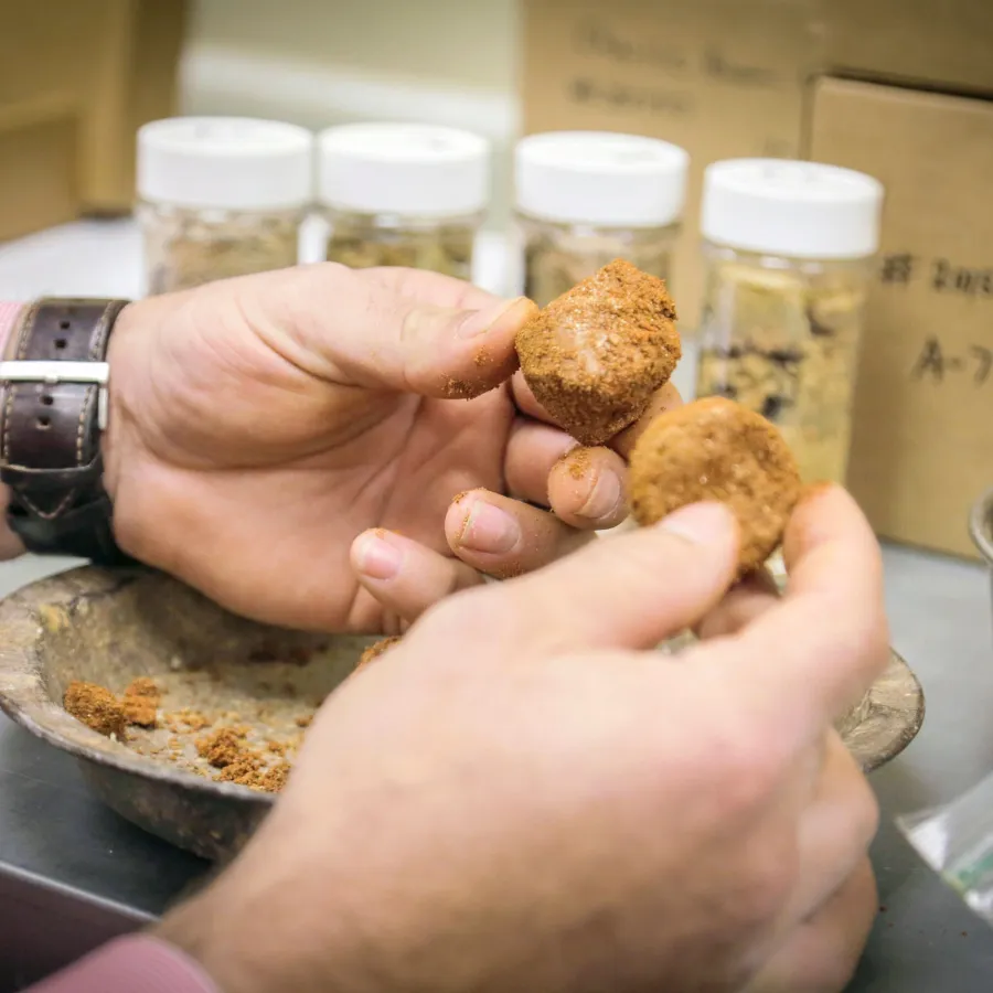 Hands holding and breaking apart brown clay or soil samples over a bowl with jars in the background.