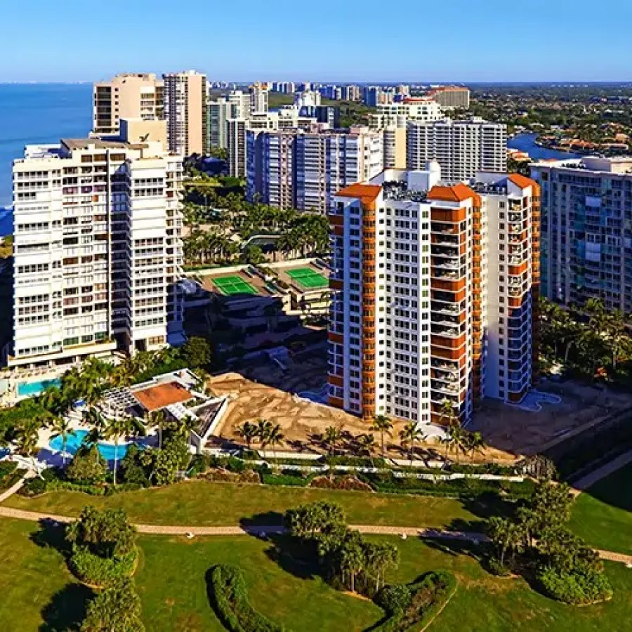Aerial view of beachfront high-rise condos with pools, palm trees, and green lawns under a clear blue sky.